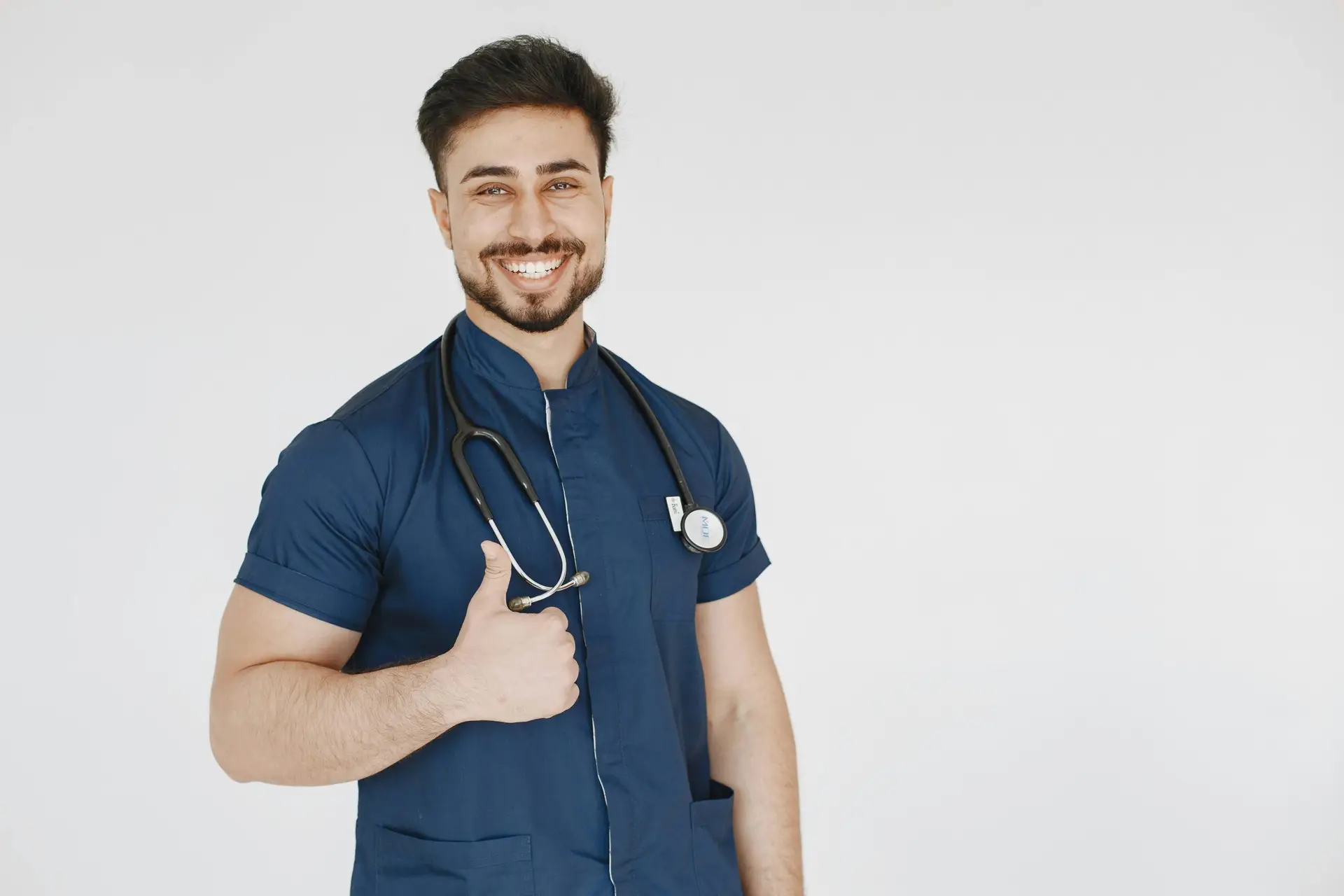 Confident male doctor in blue scrubs with a stethoscope smiling and giving a thumbs up.