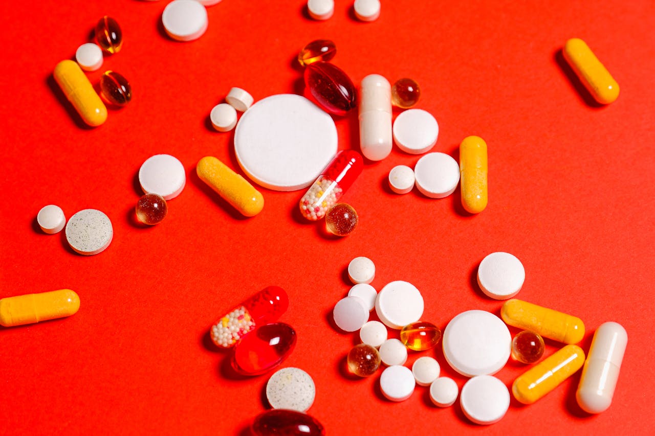 Variety of pills and capsules scattered on vibrant red backdrop, showcasing diverse medicine forms.